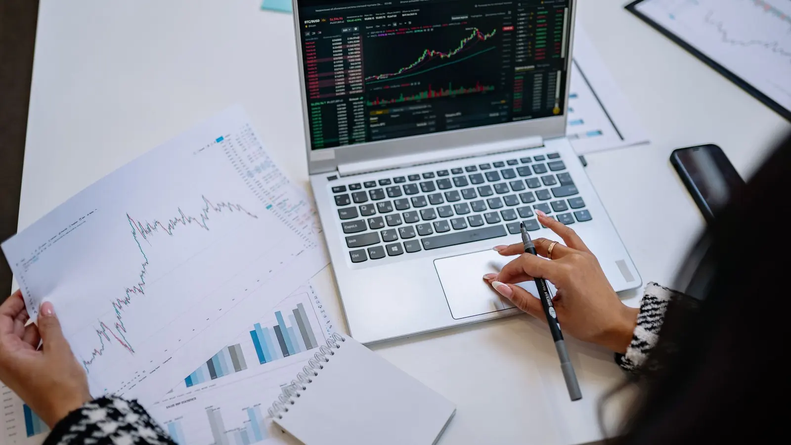 A laptop showing a price chart beside printed charts and notes