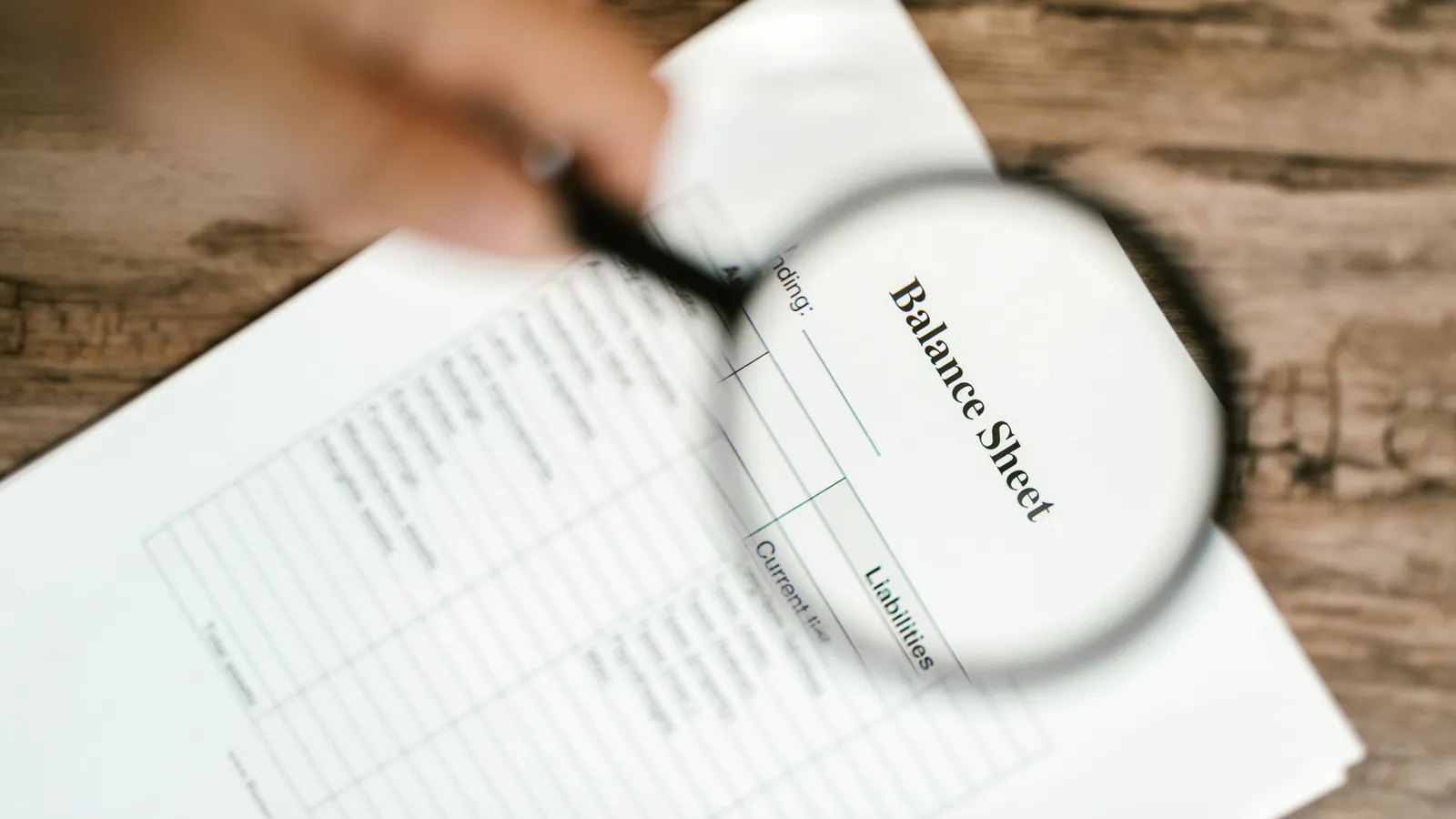 A hand holding a magnifying glass over a balance sheet