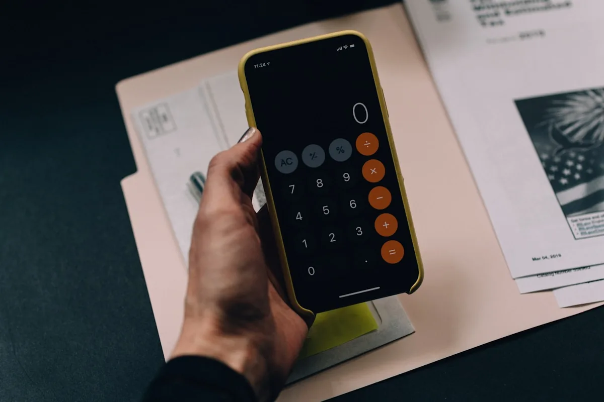 A hand holding a phone with a calculator over financial paperwork, representing the real cost of a free funded account