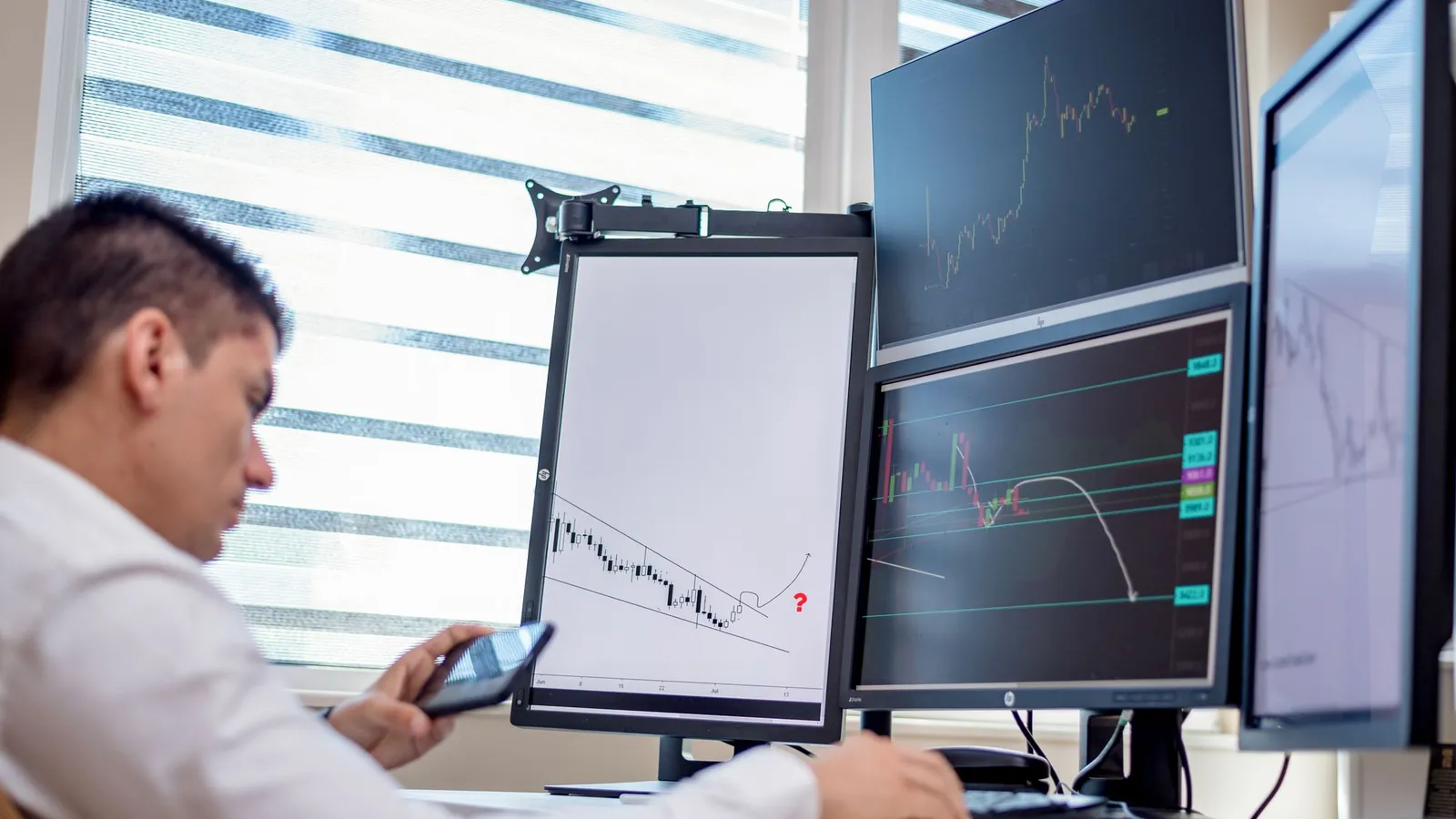 A trader checking his phone in front of screens showing charts