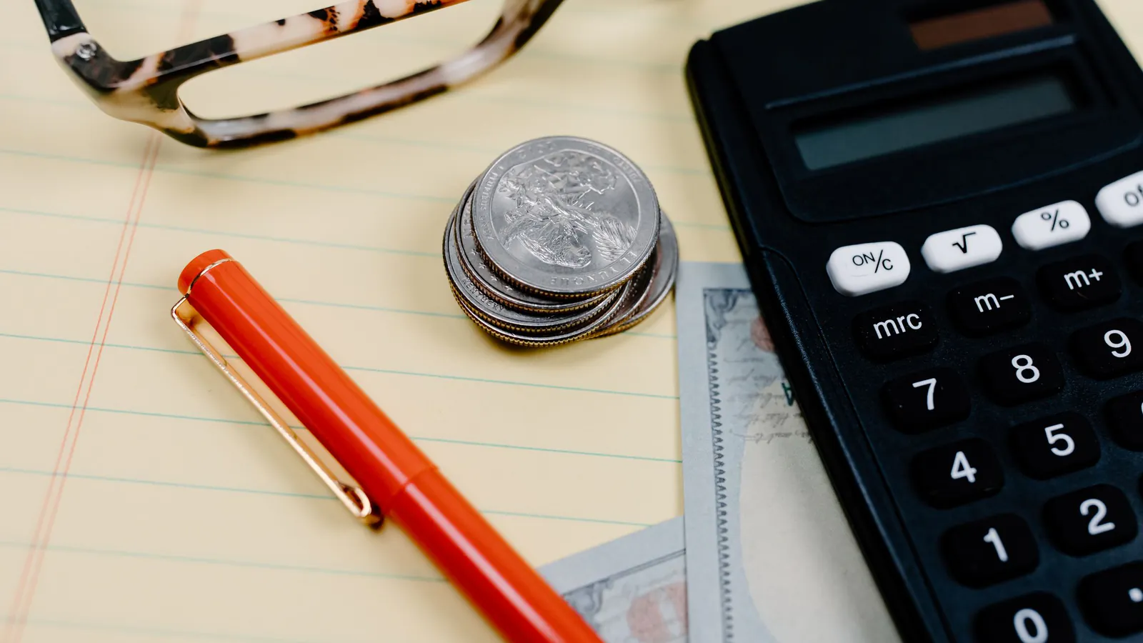 Calculator, coins, glasses and a pen on a notepad