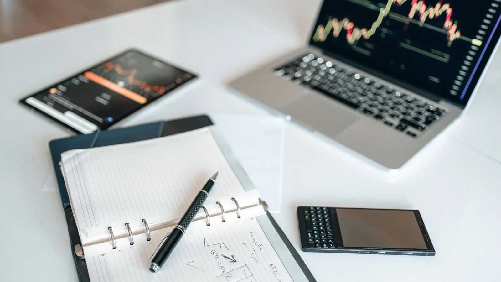 A notebook, tablet and laptop showing price charts on a white desk